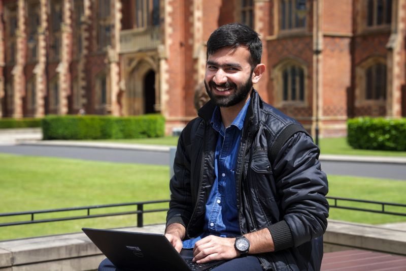 male student sat outside Queen's building on laptop